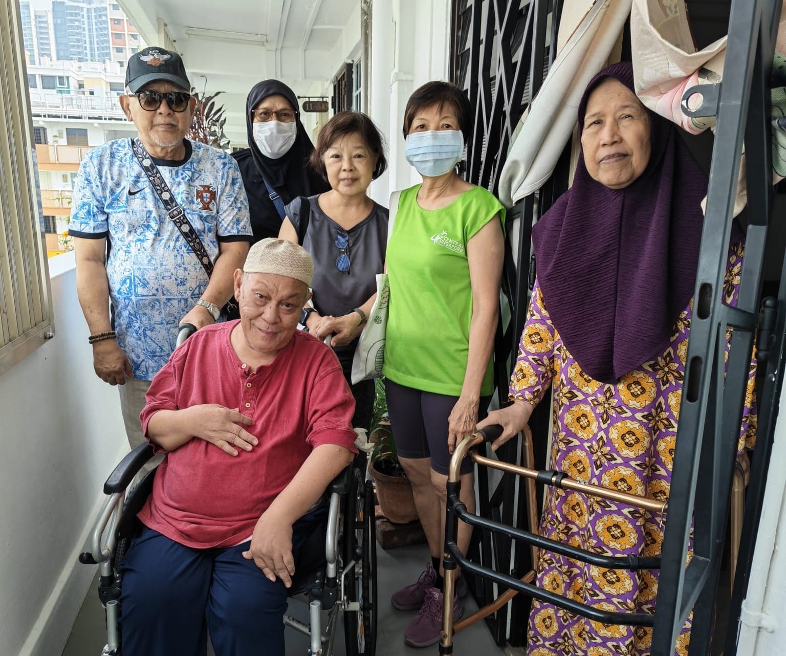 A group of six elderly residents posing together in an HDB corridor, one seated in a wheelchair and another using a walking frame.